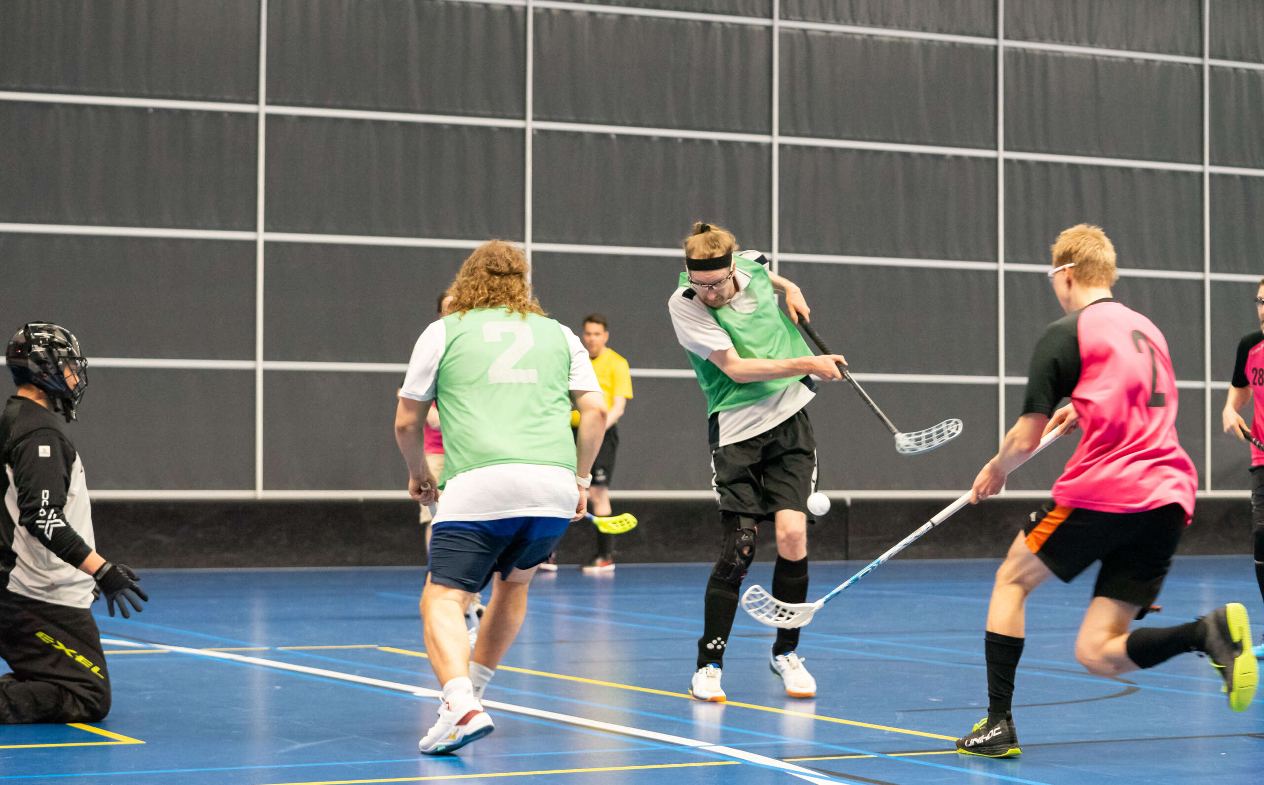 Vihreään liivin pukeutunut pelaaja syöttää joukkuekaverille maalintekopaikkaan. Vastustajajoukkueen pelaaja juoksee puolustamaan maalia.