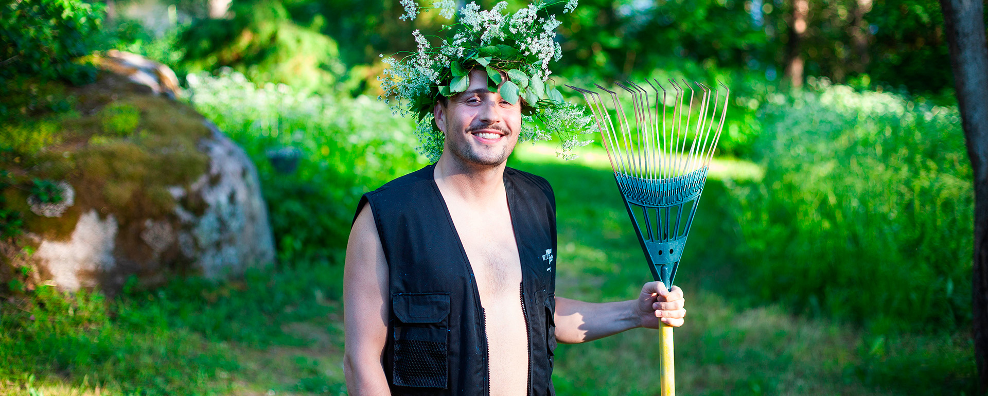 Joonas Pesonen seisoo vihreällä nurmikolla. Yllään hänellä on musta kalastusliivi, päässään kukkaseppele ja kädessään harava.