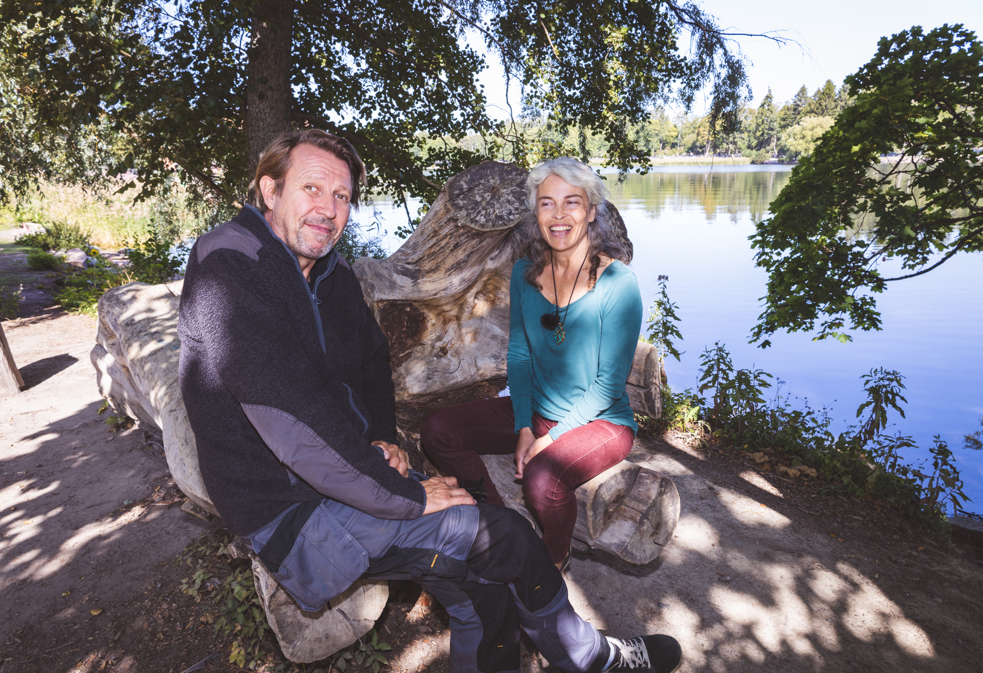 Martti Suosalo ja Irina Björklund istuvat kaatuneen puunrungon päällä, rannassa puiden alla.