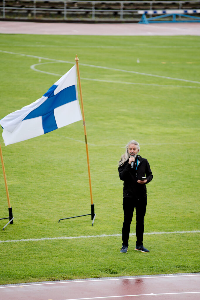 Tarmo Raatikainen puhuu mikrofoniin urheilukentän laidalla. Hänen takanaan liehuu Suomen lippu.