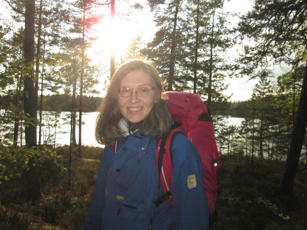 Merja Matilainen on järven rannalla metsässä retkeilyvarusteissa. Hän hymyilee.