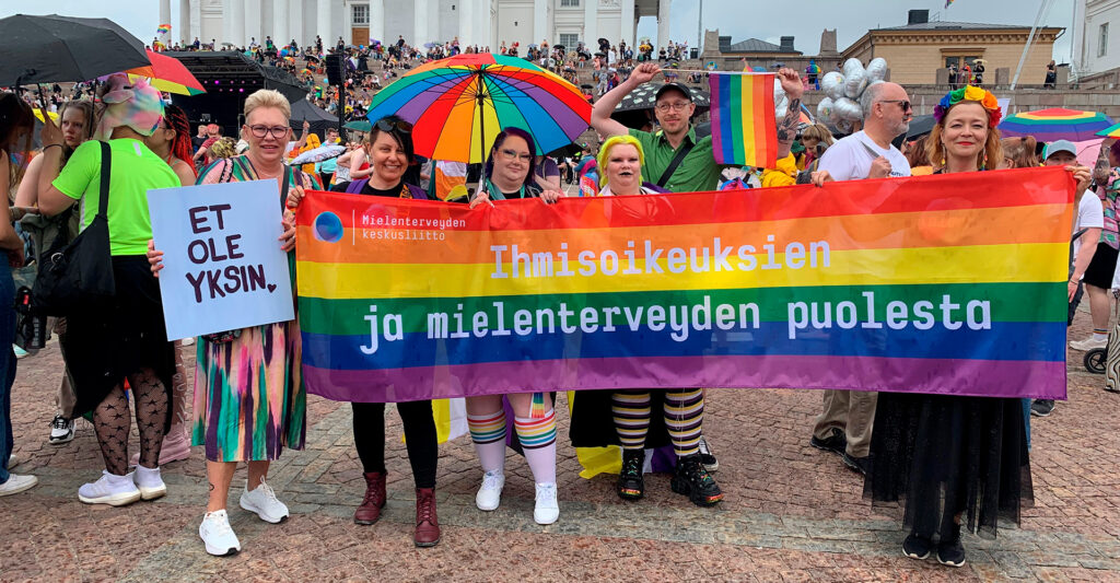 Mielenterveyden keskusliiton työntekijöitä ja vapaaehtoisia Helsingin Senaatintorilla odottamassa Pride-kulkueen alkua. Heillä on sateenkaarilippu, jossa lukee: Ihmisoikeuksien ja mielenterveyden puolesta.