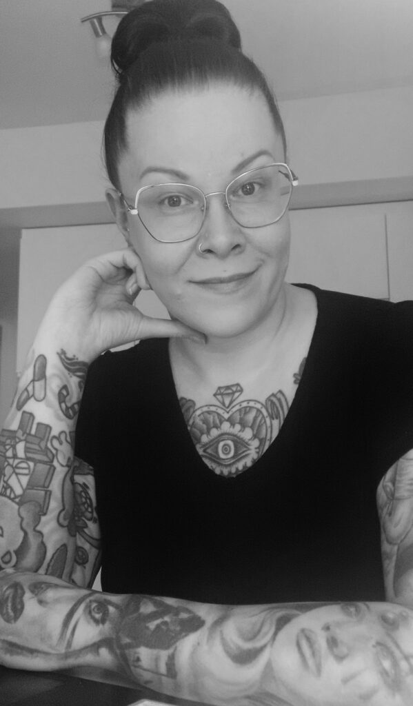 Emilia nojaa käsivarsillaan pöytään ja hymyilee. Hänellä on tatuointeja käsissään, silmälasit ja hiukset nutturalla päälaella.