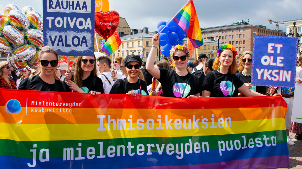 Kuusi Mielenterveyden keskusliiton työntekijää pitää kylttejä ja banderollia Helsingin Senaatintorilla ennen kulkueen starttaamista kesällä 2024. Banderolleissa lukee: "Rauhaa, toivoa, valoa" ja "Et ole yksin". Banderollissa lukee: "Ihmisoikeuksien ja mielenterveyden puolesta".