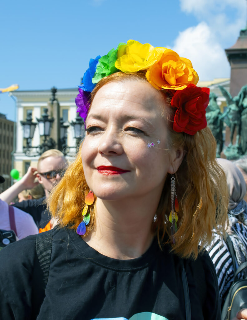 Päivi Tonteri hymyilee Helsingin Senaatintorilla, jossa näkyy pride-kulkueen osallistujia. Hänellä on värikäs kukkapanta ja sateenkaaren väriset korvakorut.