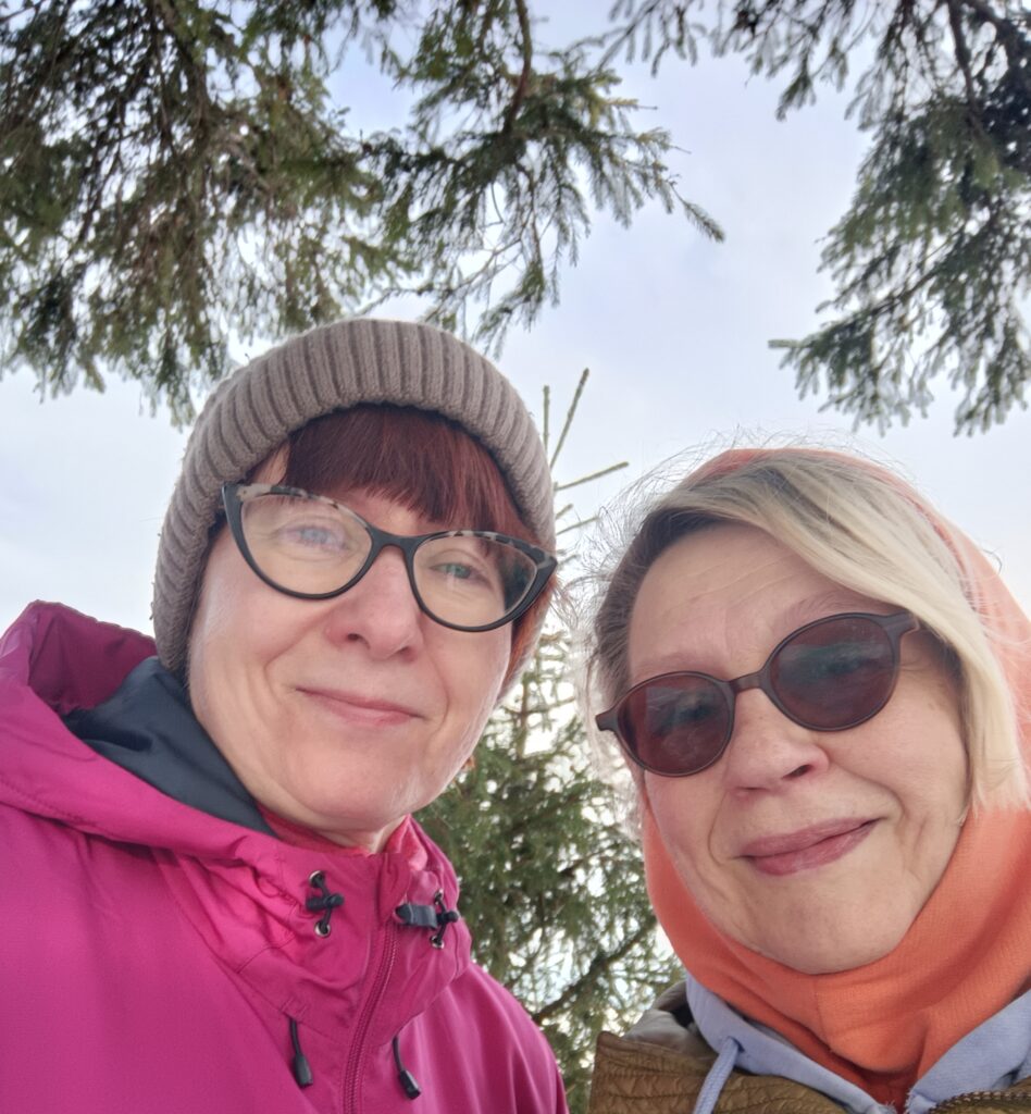 Luontolähetit Outi ja Raija hymyilevät aurinkoisena päivänä puiden siimeksessä.