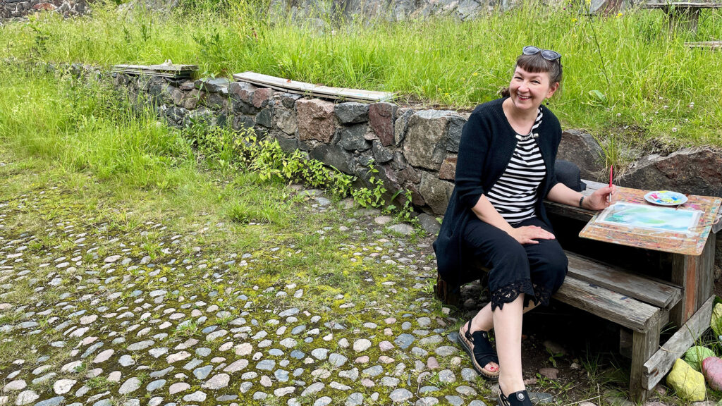 Nina Tuittu istuu Suomenlinnassa pienellä puisella penkillä ja hymyilee. Hänen edessään olevalla puisella pöydällä on paperia ja maalaustarvikkeet. Taustalla on Suomenlinnan linnoituksen kivimuuria.