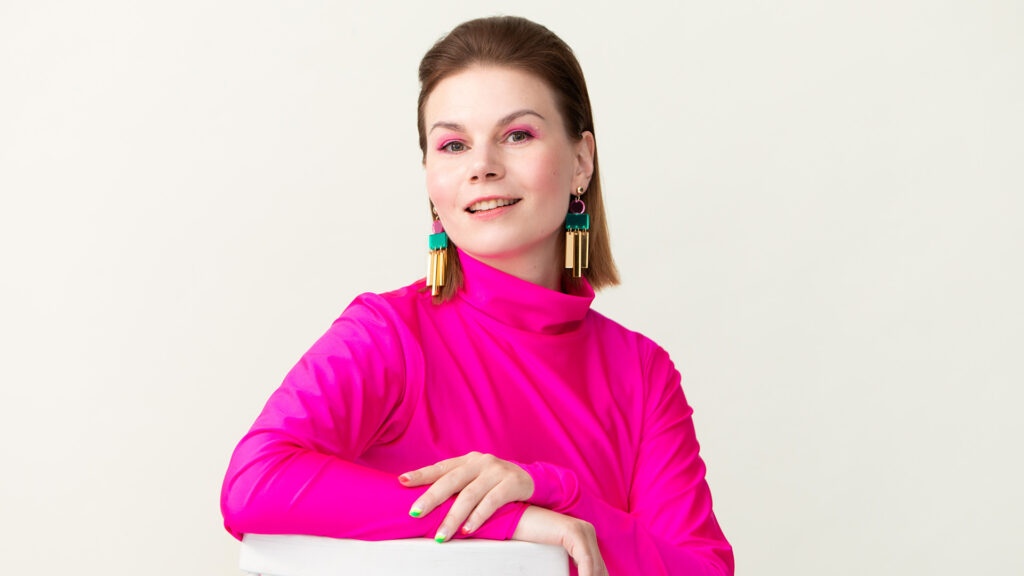 Emilia Kujala nojaa käsivarrellaan tuolin selkänojaan ja hymyilee. Hänellä on pinkki paita, isot korvakorut ja tumma olkapäille ulottuva tukka.