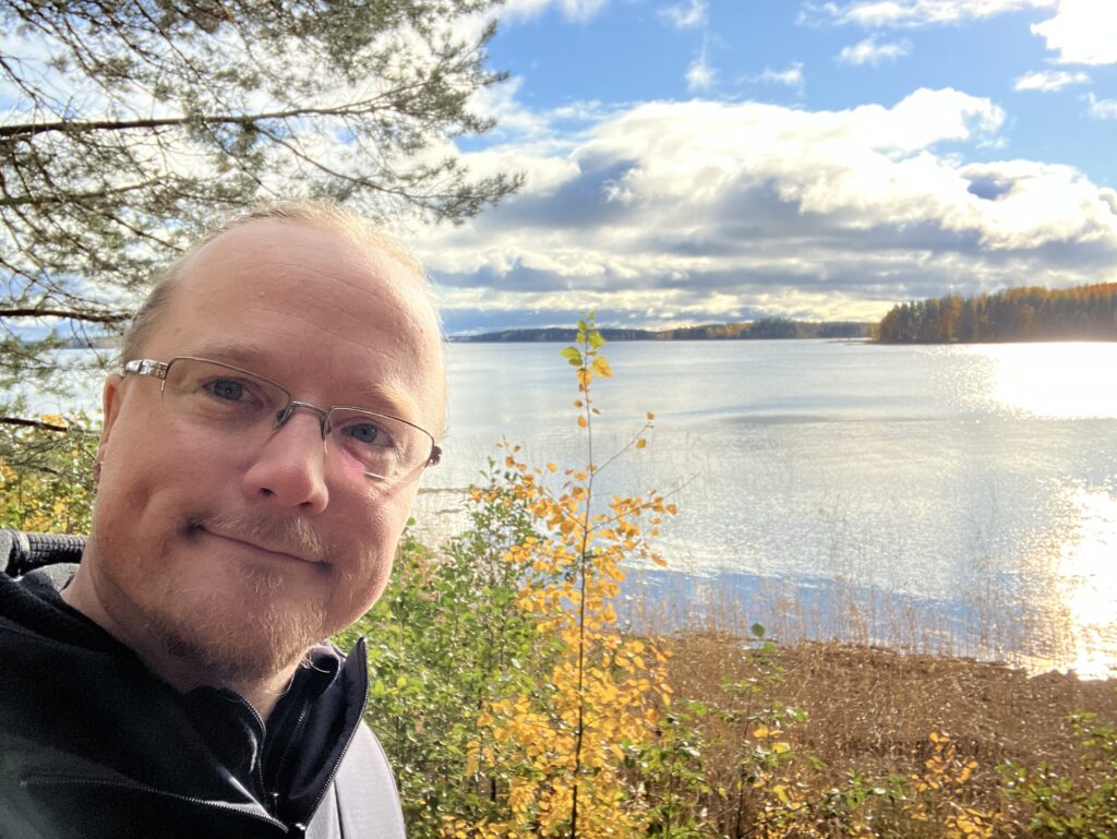 Esko Poutanen hymyilee. Hänen takanaan on järven ranta ja puissa keltaisia lehtiä.