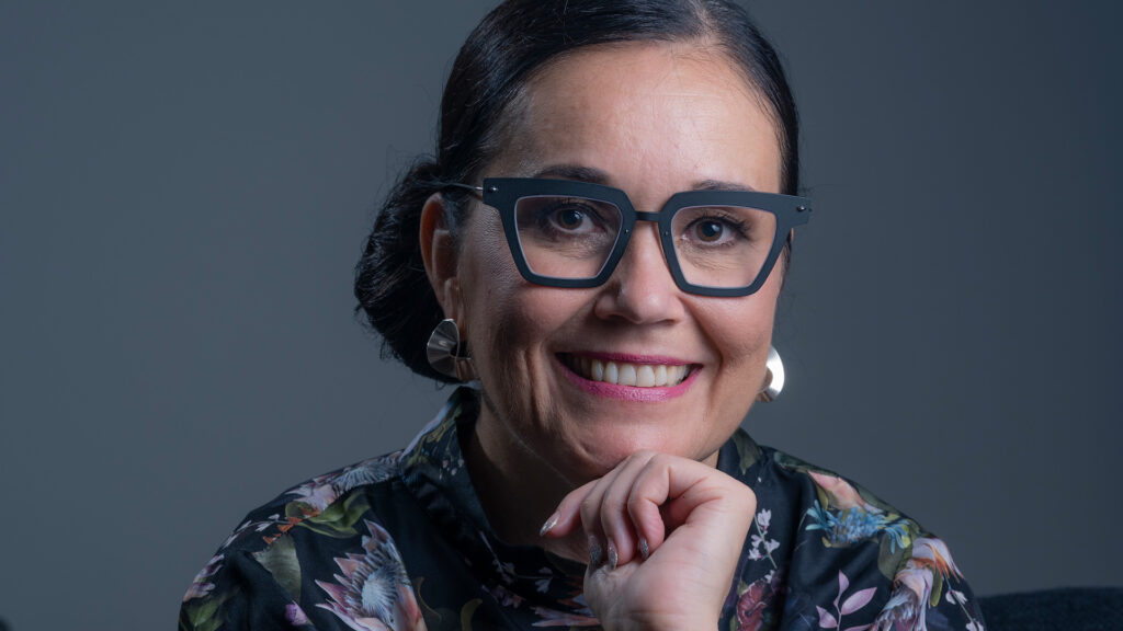 Nina Lyytinen nojaa leukaa kämmeneensä ja hymyilee. Hänellä on tummat hiukset nutturalla niskassa ja silmälasit.