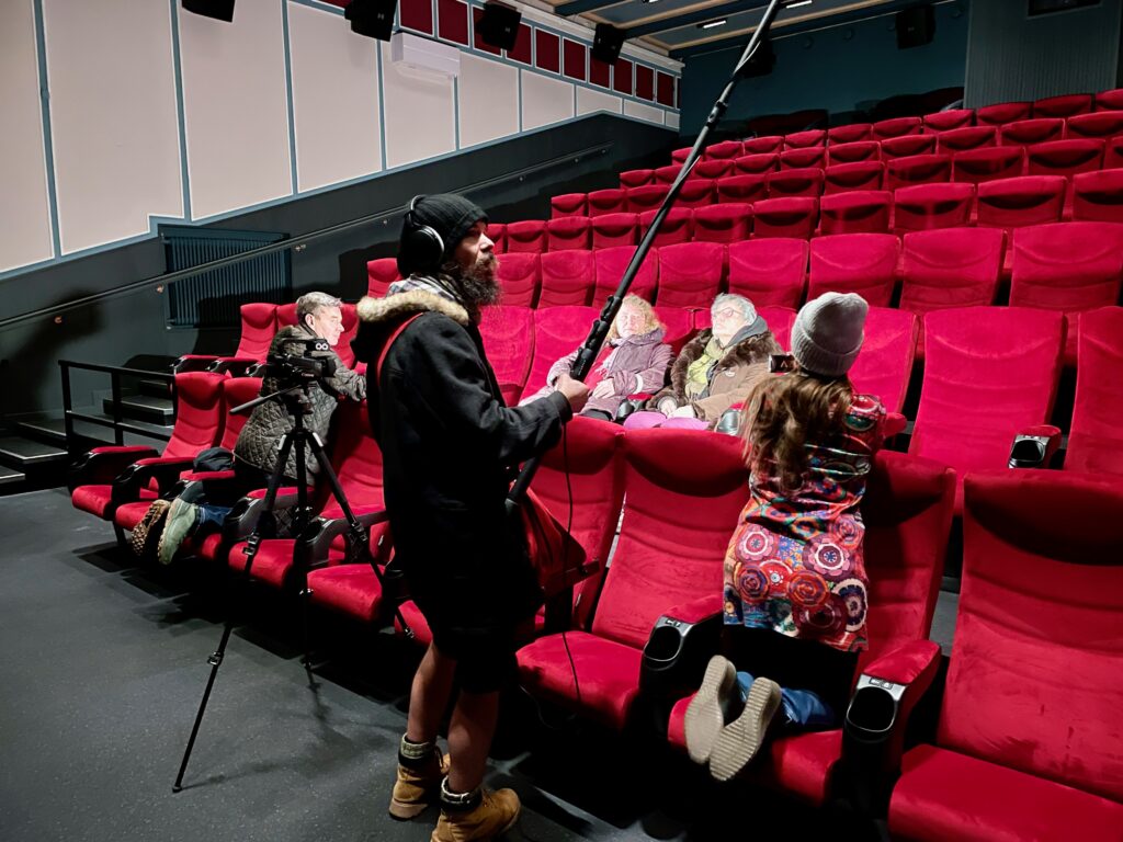 Kaksi ihmistä istuu elokuvateatterin punaisilla penkeillä. Heidän edessään on kamera ja äänittäjä, joka kannattelee mikrofonia. Kaksi ihmistä on polvillaan elokuvateatterin eturivissä selin valkokankaalle.