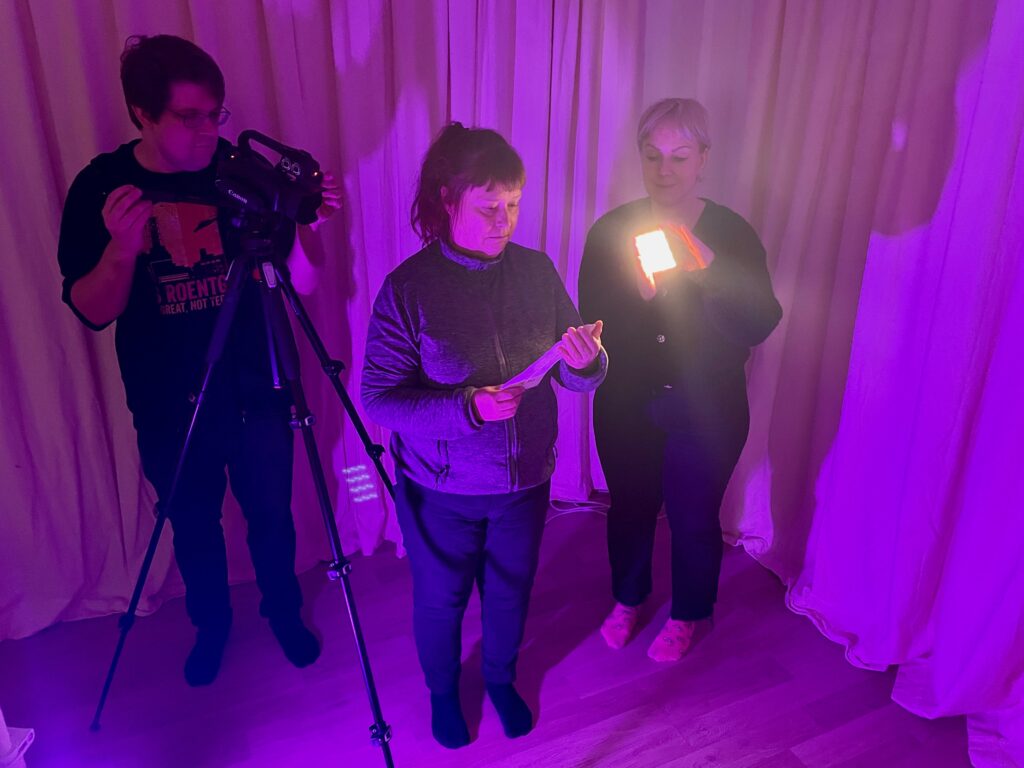 Kolme ihmistä seisoo violetiksi valaistussa huoneessa. Yksi käyttää videokameraa, toinen pitää kohdevaloa ja kolmas lukee paperista.