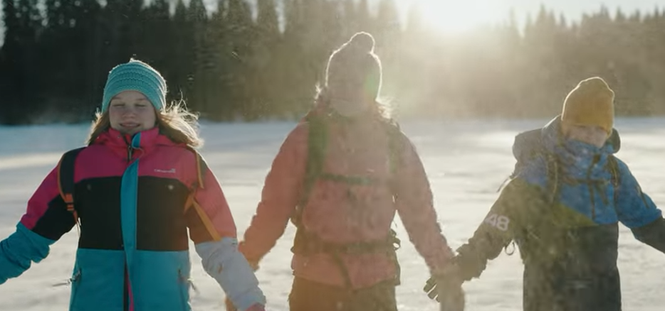 Kolme lasta pitää toisiaan kädestä kiinni talvimaisemassa.