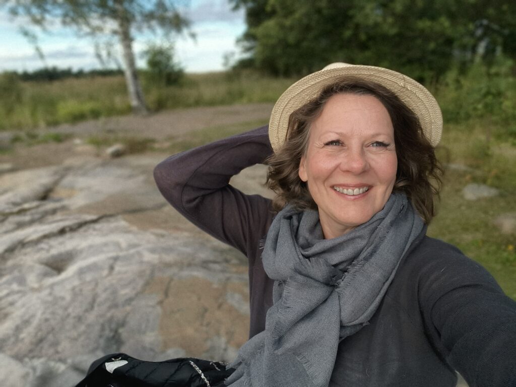 Krista Launonen istuu kalliolla ja pitää kiinni lierihatustaan. Hän hymyilee.
