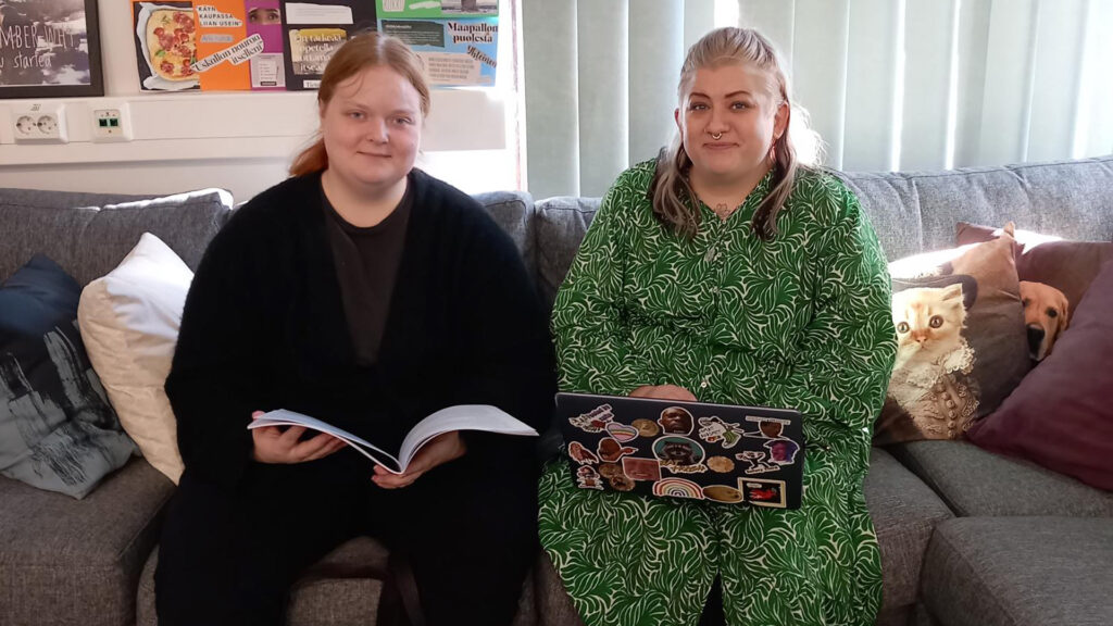 Sara ja Jenni istuvat Omat Avaimet 4You -toiminnan tiloissa sohvalla. Toisella on kädessään vihko ja toisella sylissään tietokone. Molemmat hymyilevät.