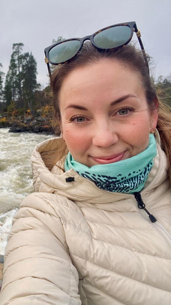 Sanna hymyilee kuohuvan kosken rannalla. Yllään hänellä on kevytuntuvatakki ja aurinkolasit on nostettu otsalle.