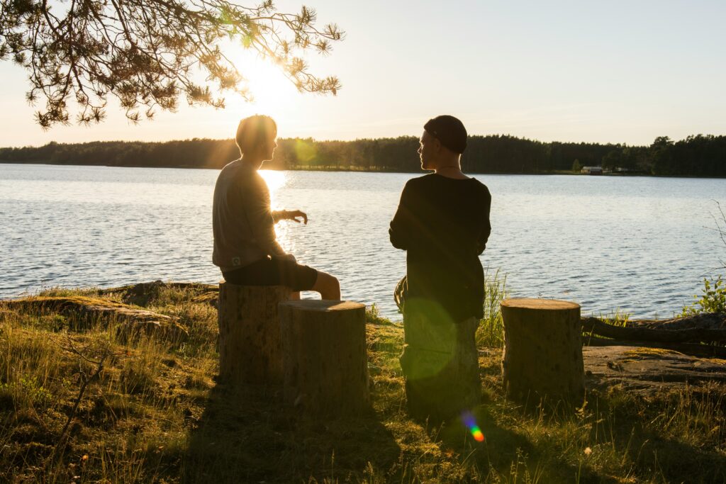 Kaksi ihmistä istuu puupölleillä järven rannalla auringon laskiessa.
