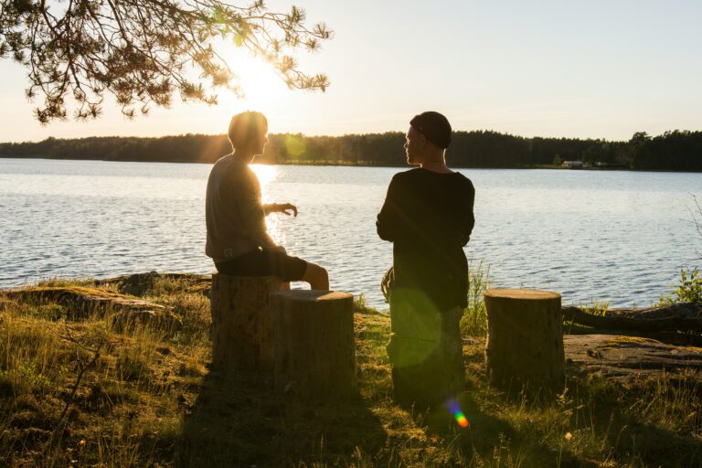 Kaksi ihmistä istuu puupölleillä järven rannalla auringon laskiessa.